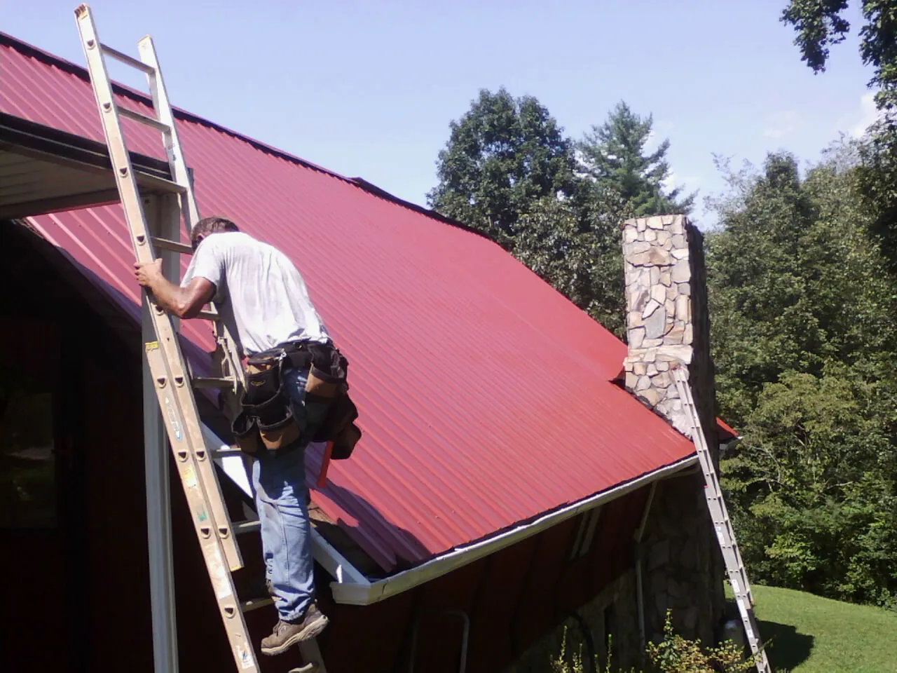 Roofer on a ladder beside a red metal roof and stone chimney — Soffit & Fascia Repair by Roofing Brothers in Defiance
